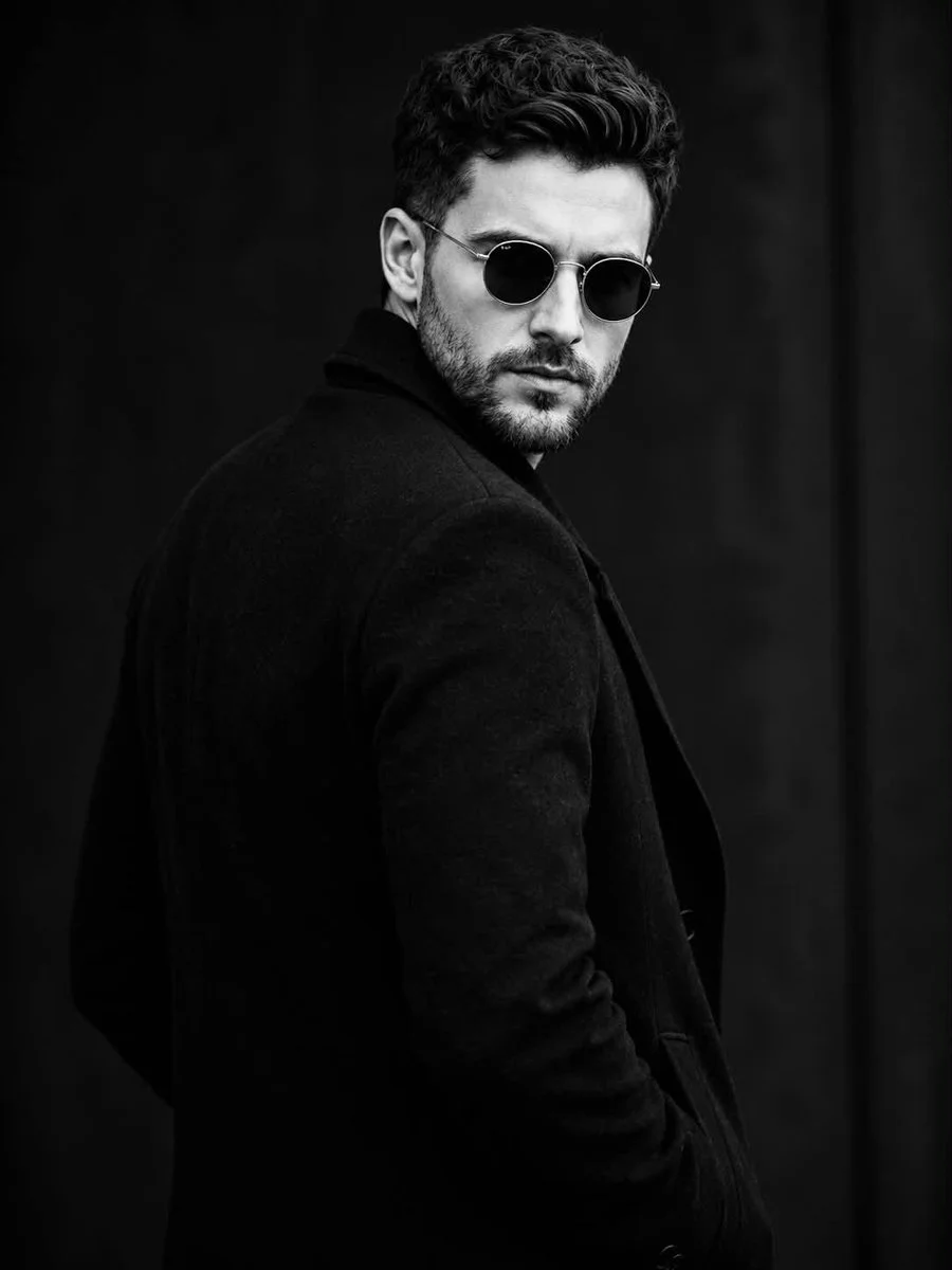 A bearded man in sunglasses looks back from a dark studio.