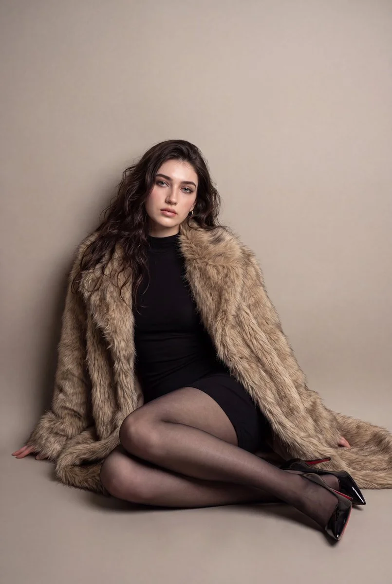 A model sits on the floor in a warm studio wearing a fur coat.