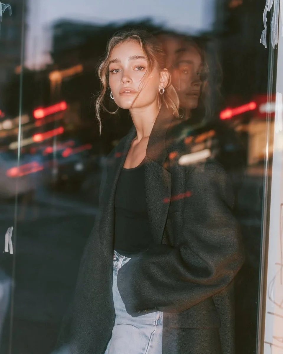 A stylish woman in charcoal blazer poses by glass storefront at blue hour.