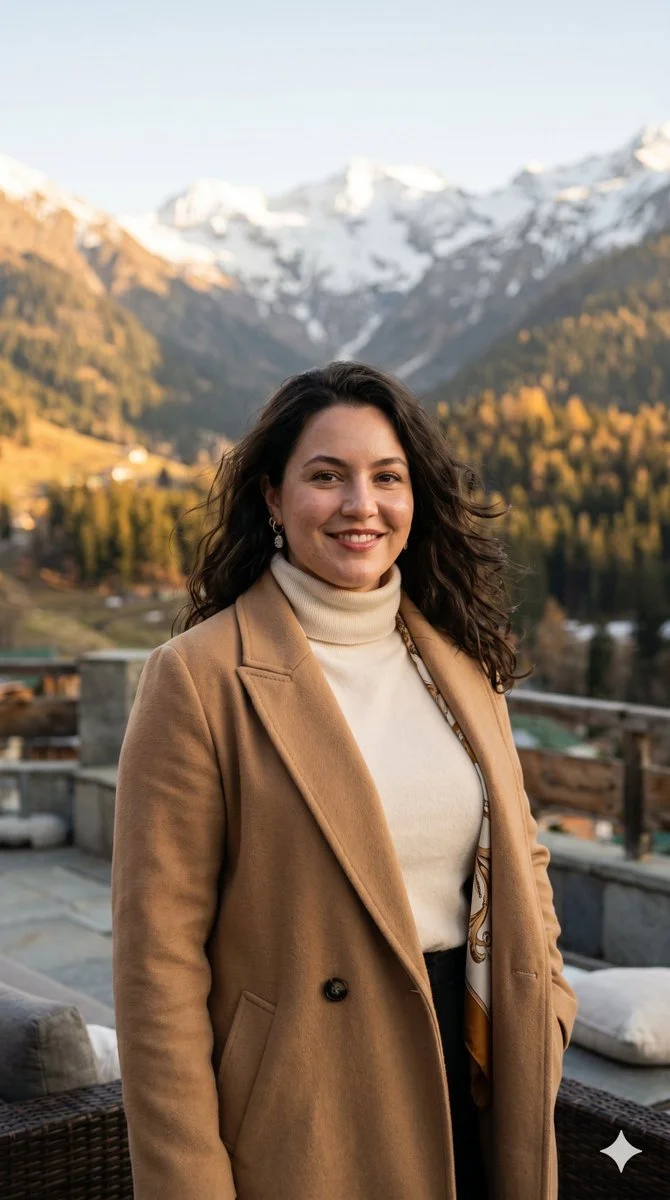 A woman in a camel coat stands on a mountain terrace at golden hour
