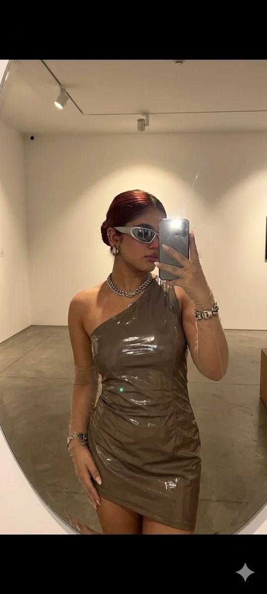 A woman in a futuristic dress takes a mirror selfie in a gallery.