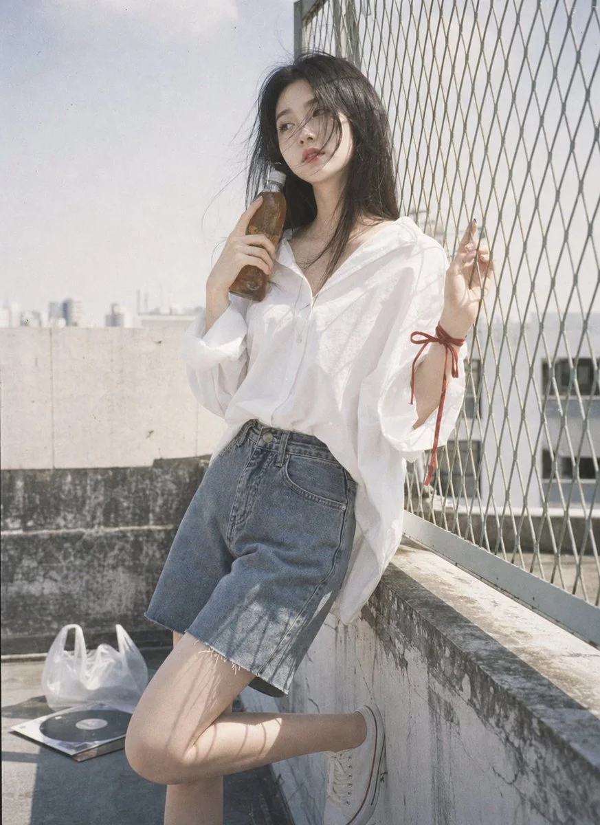 A woman in white leans by a fence with a glass bottle