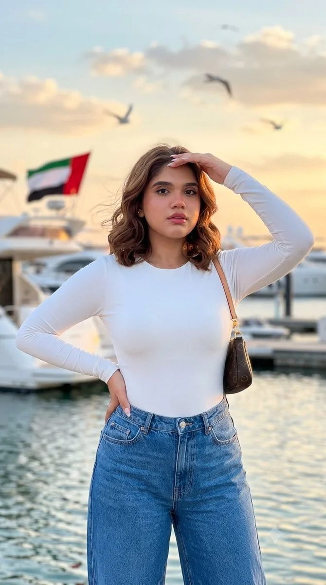 A woman in white top and jeans poses by the marina at sunset