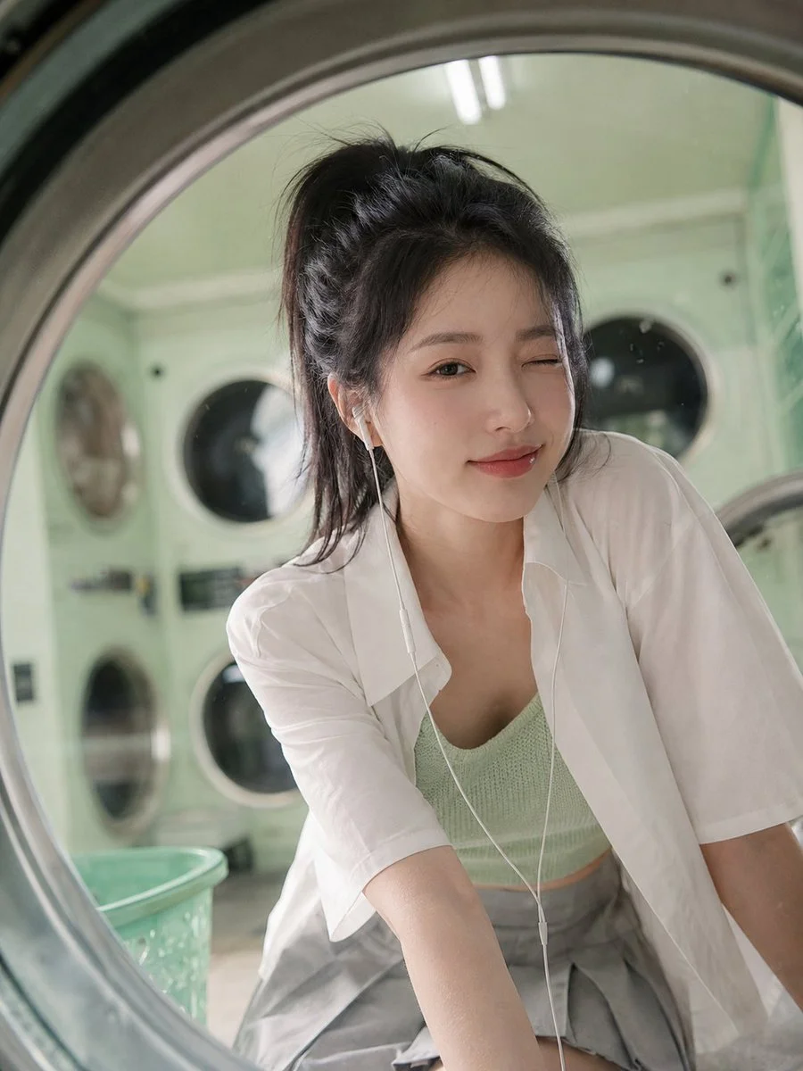 A woman peeks through a washing machine window in a mint green laundromat.