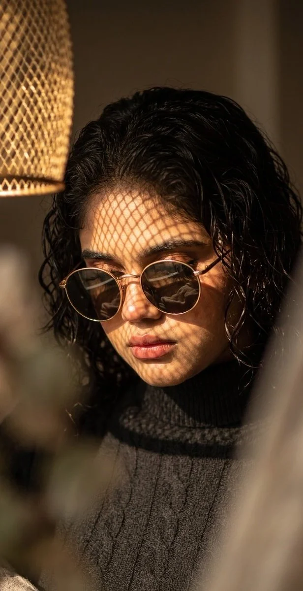 A woman with gold sunglasses gazes downward in warm dappled sunlight.