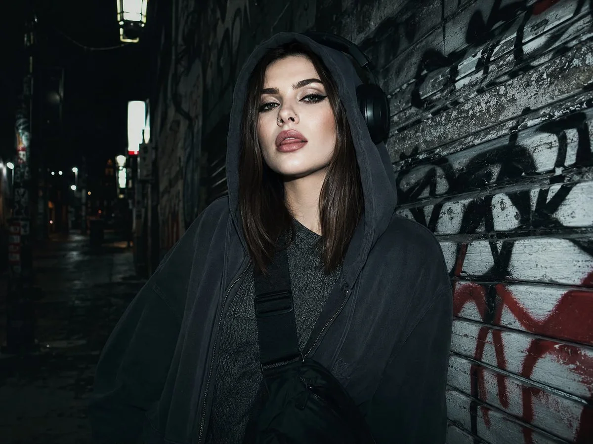 A young woman in an alley poses with headphones and a hoodie