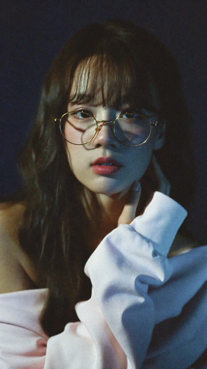 A young woman in gold glasses touches her cheek under dim light