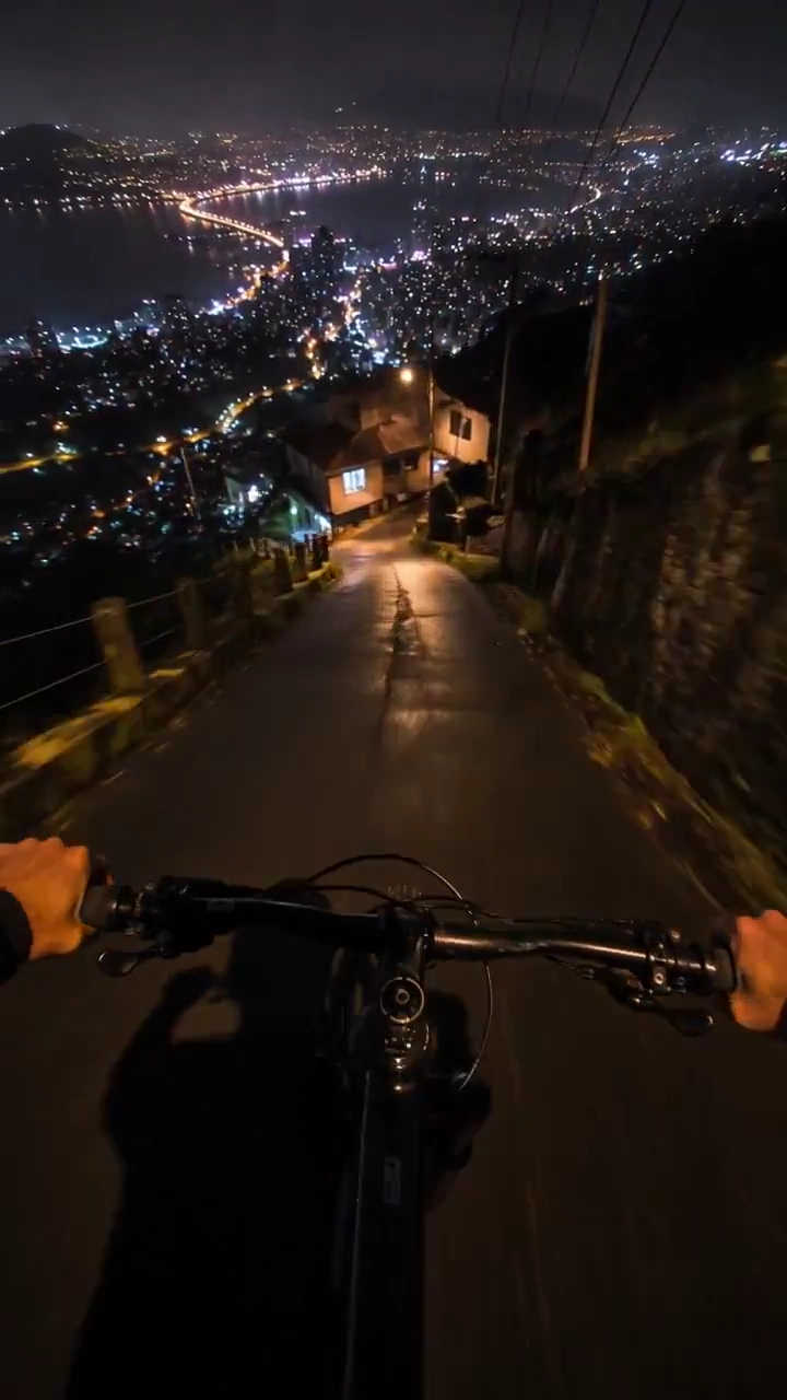 Handlebar View Descends Steep Wet Road Above Glowing City