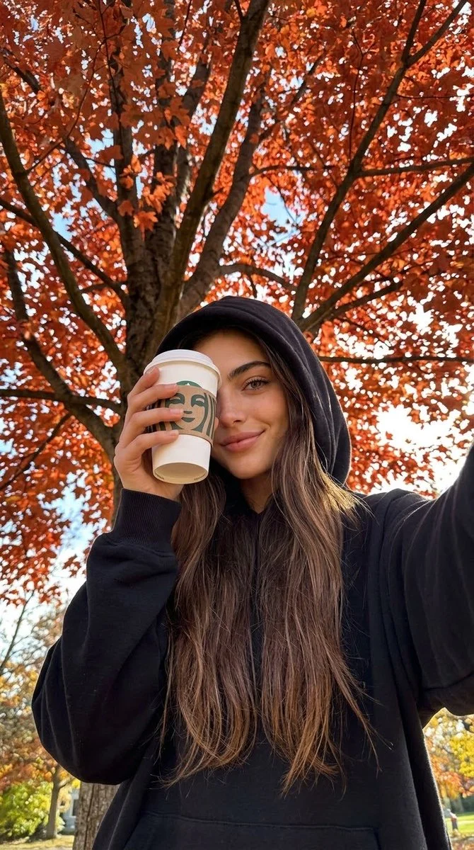 Girl in black hoodie holds coffee under autumn tree