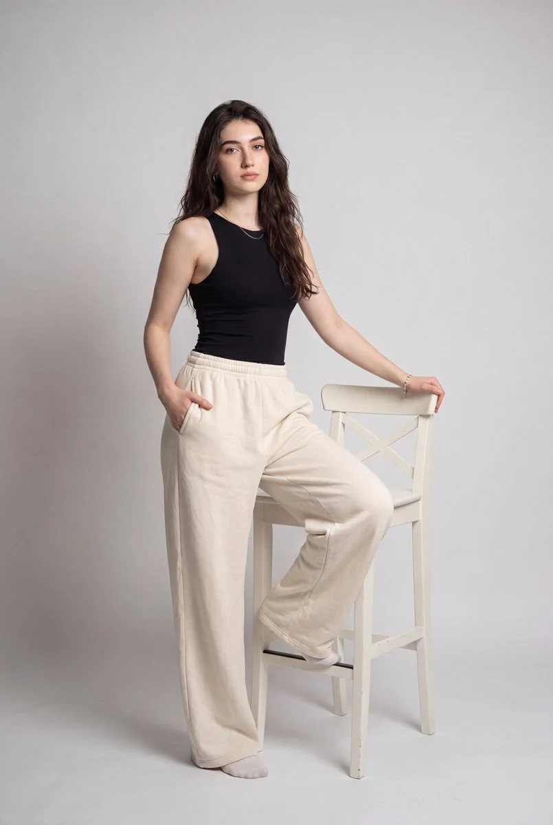 Model in black tank and cream pants poses with one leg on chair