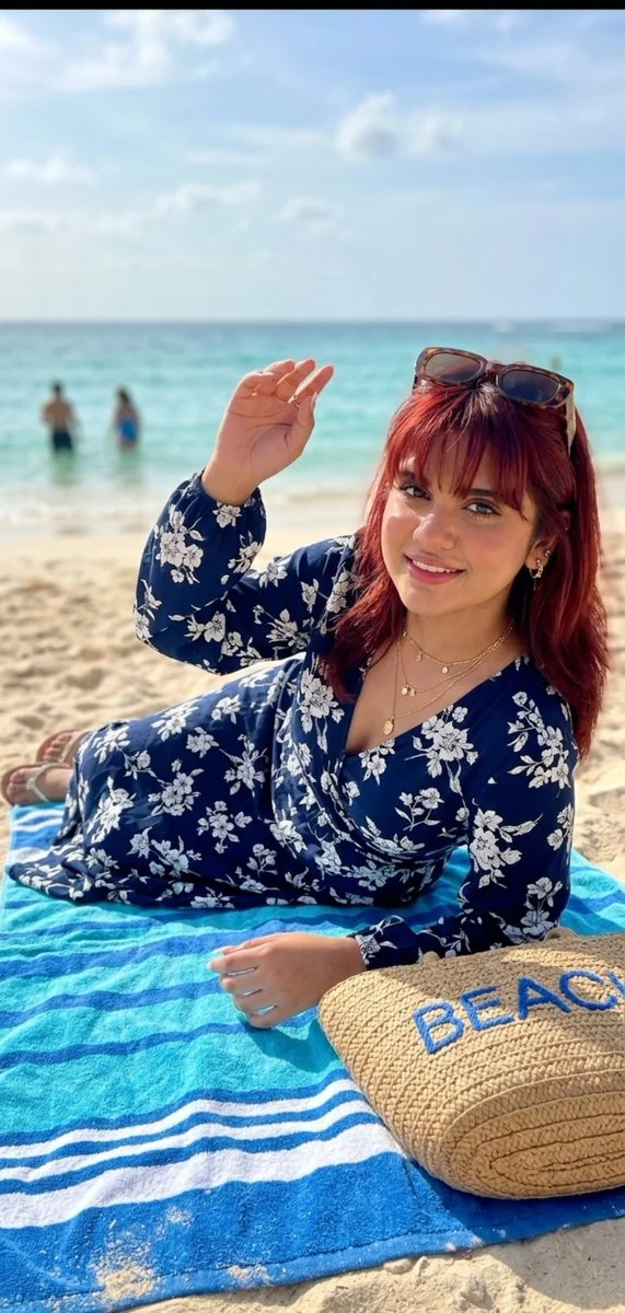 Woman with floral crop top smiles beside straw bag on beach