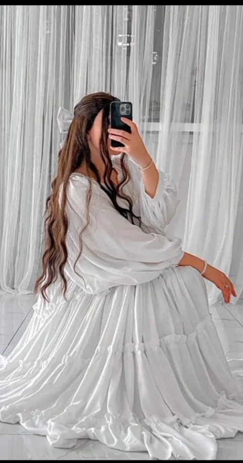 Young woman in white satin dress captures mirror selfie on marble floor.